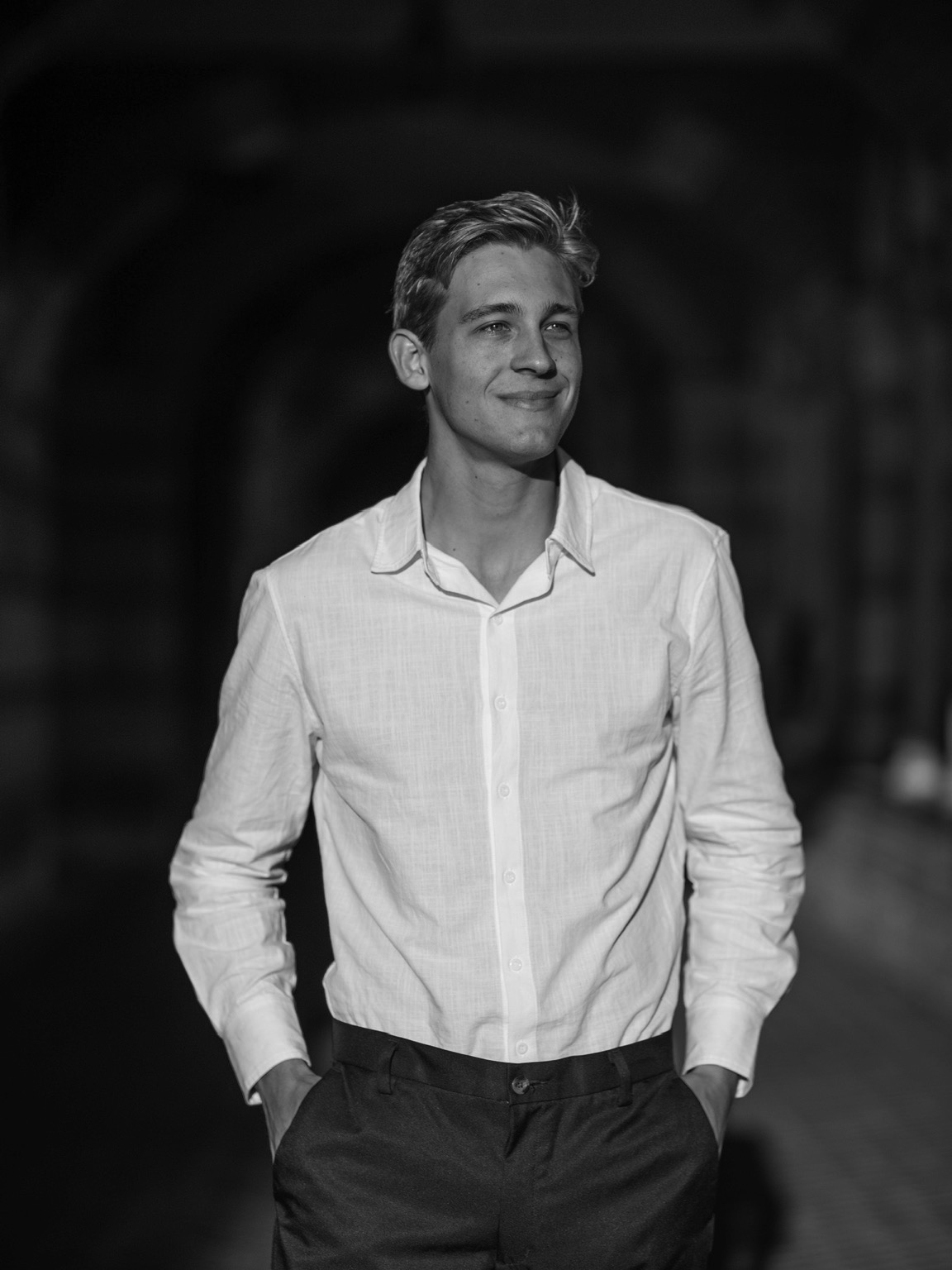 Alexander in a white button-down, hands in pockets, smiling under brick archways